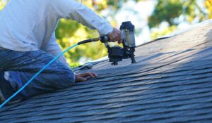 A worker examines shingles on a residential roof under bright sky, with trees visible in the background.