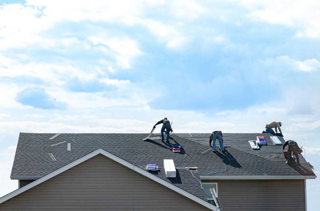 Roofing workers inspect exposed roof decking beside removed shingles on a residential home.